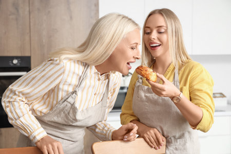 Beautiful young woman and her mother with eating tasty bun in kitchenの写真素材