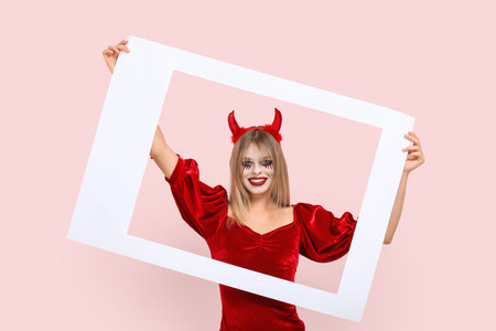 Young woman dressed for Halloween as devil with frame on pink background