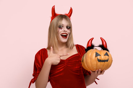 Young woman dressed for Halloween as devil pointing at pumpkin on pink backgroundの写真素材