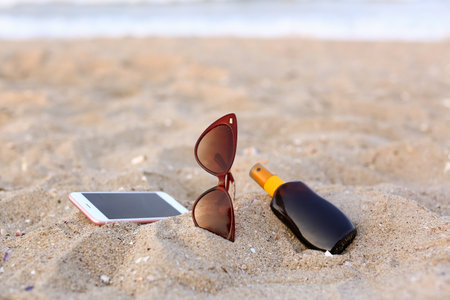 Stylish sunglasses with sunscreen and phone on sandの写真素材
