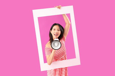 Young beautiful Asian woman with frame and megaphone on purple backgroundの写真素材