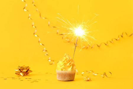 Tasty birthday cupcake with sparkler and decor on yellow backgroundの写真素材
