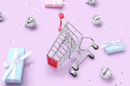Composition with small shopping cart, gift boxes and Christmas balls on purple background, closeupの写真素材