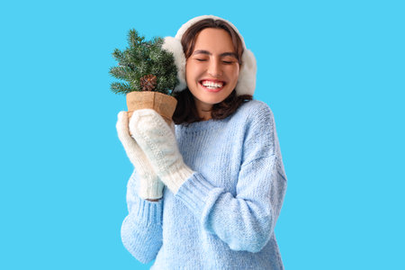 Young woman in warm clothes with small Christmas tree on blue backgroundの写真素材
