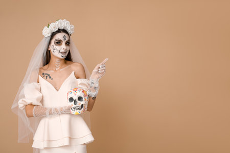 Young woman dressed as dead bride for Halloween party with painted human skull pointing at something on beige backgroundの写真素材