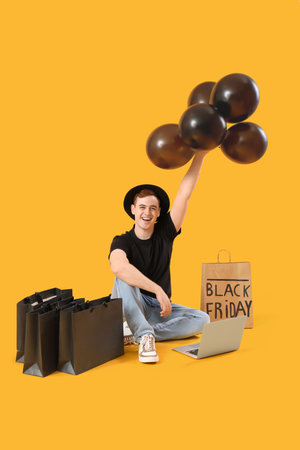 Happy young man with shopping bags, balloons and laptop sitting on yellow background. Black Friday saleの写真素材