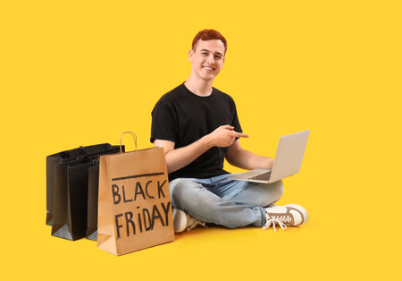 Happy young redhead man with shopping bags pointing at laptop sitting on yellow background. Black Friday saleの写真素材