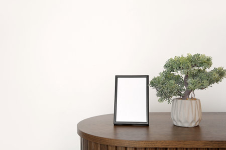 Blank frame and decorative tree in pot on wooden cabinet near white wall, closeup