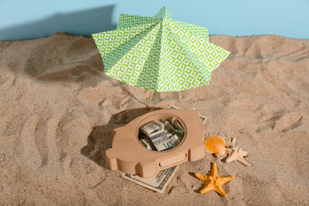 Wooden piggy bank with money and mini umbrella on sand near blue wall. Concept of savings for travelの写真素材
