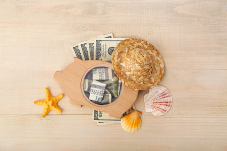 Piggy bank with money, wicker hat and seashells on beige wooden background. Concept of savings for...