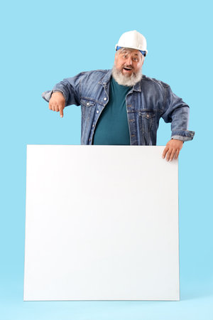 Portrait of senior man in hardhat pointing at blank poster on blue background. Labor Day celebrationの写真素材