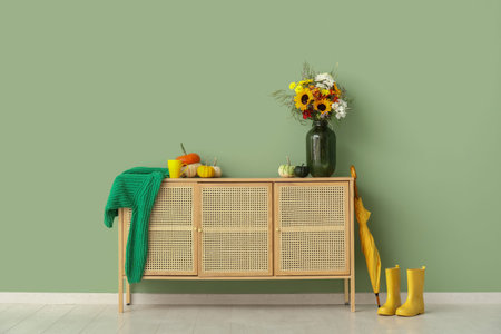 Wooden cabinet with bouquet of autumn flowers, pumpkins, umbrella and rubber boots near green wall