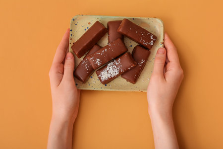 Woman holding plate with tasty chocolate covered coconut candies on orange background