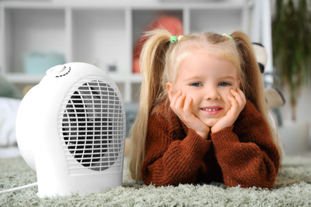 Cute little girl warming near electric fan heater in bedroomの写真素材