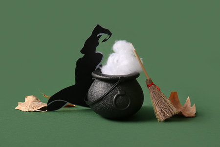 Halloween composition with cauldron, broom, leaves and witch hat on green background