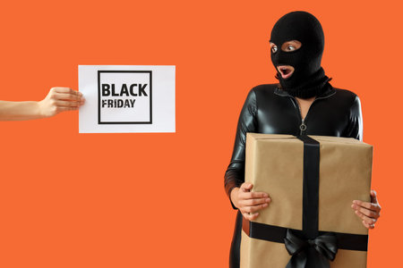 Young woman in balaclava with gift on orange background. Black Friday sale