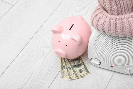 Electric fan heater, piggy bank, money and hat on floor. Heating seasonの写真素材