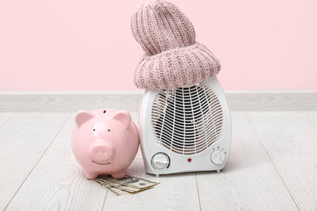Electric fan heater, piggy bank, money and hat on floor. Heating seasonの写真素材