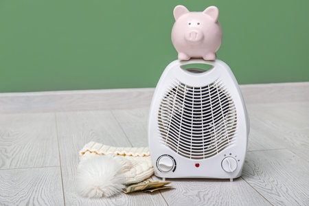 Electric fan heater, piggy bank, money and hat on floor. Heating seasonの写真素材