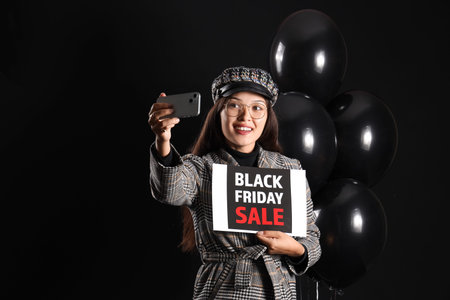 Young Asian woman with card and balloons taking selfie on dark background. Black Friday saleの写真素材