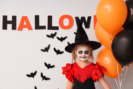 Little girl dressed for Halloween as witch with balloons and decor near light wallの写真素材