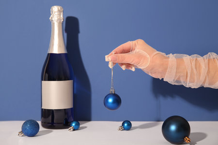 Female hand with Christmas balls and bottle of champagne on white table against blue backgroundの写真素材