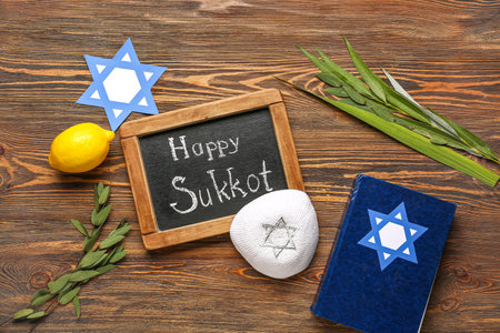 Four species (lulav, hadas, arava, etrog), paper David star, kippah and chalkboard with text HAPPY SUKKOT on wooden backgroundの写真素材