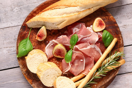 Plate with slices of tasty prosciutto, basil, bread, Italian Grissini and figs on gray wooden backgroundの写真素材