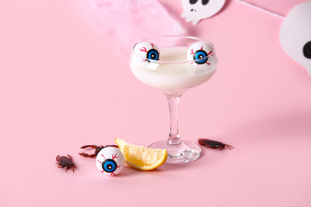 Glass of delicious white cocktail, lemon piece, eyes and candy bugs for Halloween celebration on pink backgroundの写真素材