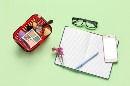 Shopping basket with makeup cosmetics, modern mobile phone, eyeglasses and notebook on green backgroundの写真素材
