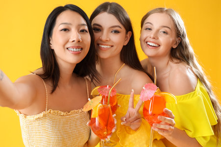 Beautiful young women with glasses of tasty spritz cocktail taking selfie on yellow backgroundの写真素材