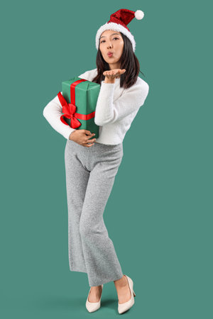 Young beautiful Asian woman in Santa hat with gift box blowing kiss on green backgroundの写真素材