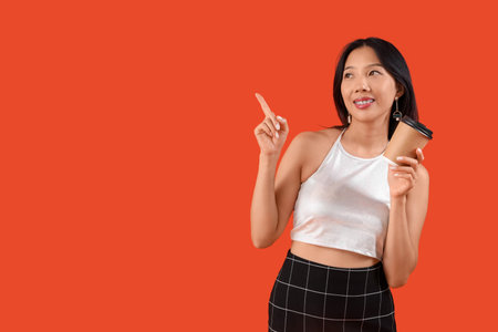 Happy young beautiful Asian woman with cup of coffee pointing at something on orange backgroundの写真素材