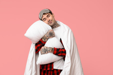Tattooed young man in pajamas with pillow and blanket on pink backgroundの写真素材