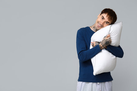 Tattooed young man in pajamas with pillow on gray backgroundの写真素材