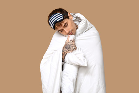 Tattooed young man in pajamas with blanket sleeping on brown backgroundの写真素材