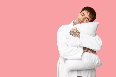 Tattooed young man in pajamas with pillow on pink backgroundの写真素材