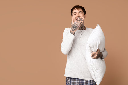 Tattooed young man in pajamas with pillow on brown backgroundの写真素材