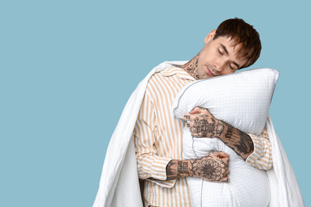 Tattooed young man in pajamas with pillow and blanket on blue backgroundの写真素材