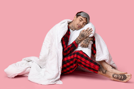 Tattooed young man in pajamas with pillow and blanket sitting on pink backgroundの写真素材