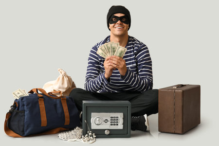 Male thief with dollar banknotes and stolen goods on gray background