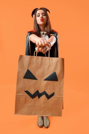 Young woman dressed for Halloween with gift bag blowing kiss on orange backgroundの写真素材