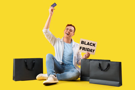 Young man with credit card and shopping bags on yellow background. Black Friday sale