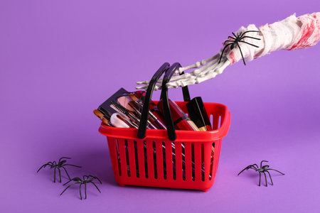 Skeleton hand with shopping basket of different decorative cosmetics and Halloween decor on purple backgroundの写真素材