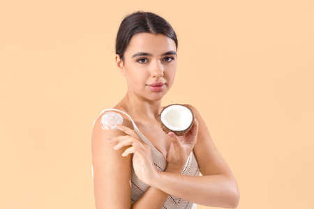 Beautiful young woman applying coconut oil on her body against beige backgroundの写真素材