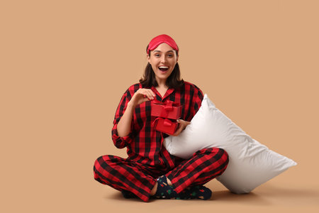 Beautiful young woman in pajamas with pillow and gift boxes sitting on brown backgroundの写真素材
