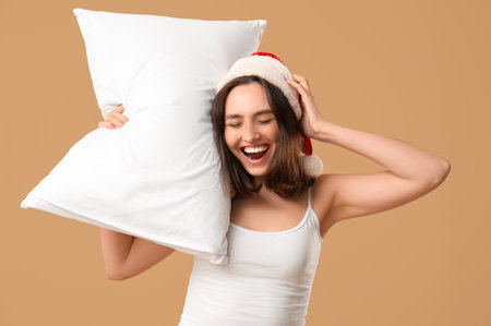 Beautiful young woman in pajamas and Santa hat with pillow on brown backgroundの写真素材