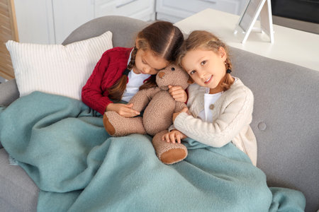 Cute little girls with toy bear and warm plaid sitting on sofa at homeの写真素材