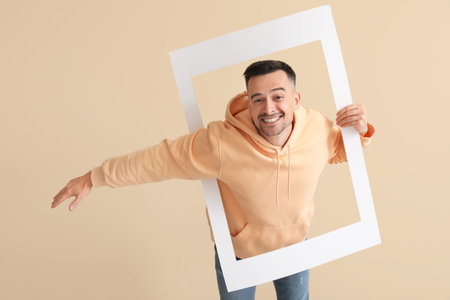 Happy young man with frame on beige background