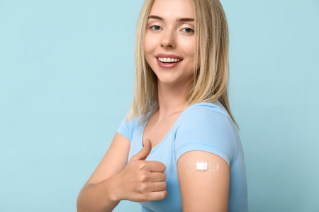 Young woman with medical patch on arm showing thumb-up against blue background. Vaccination concept
